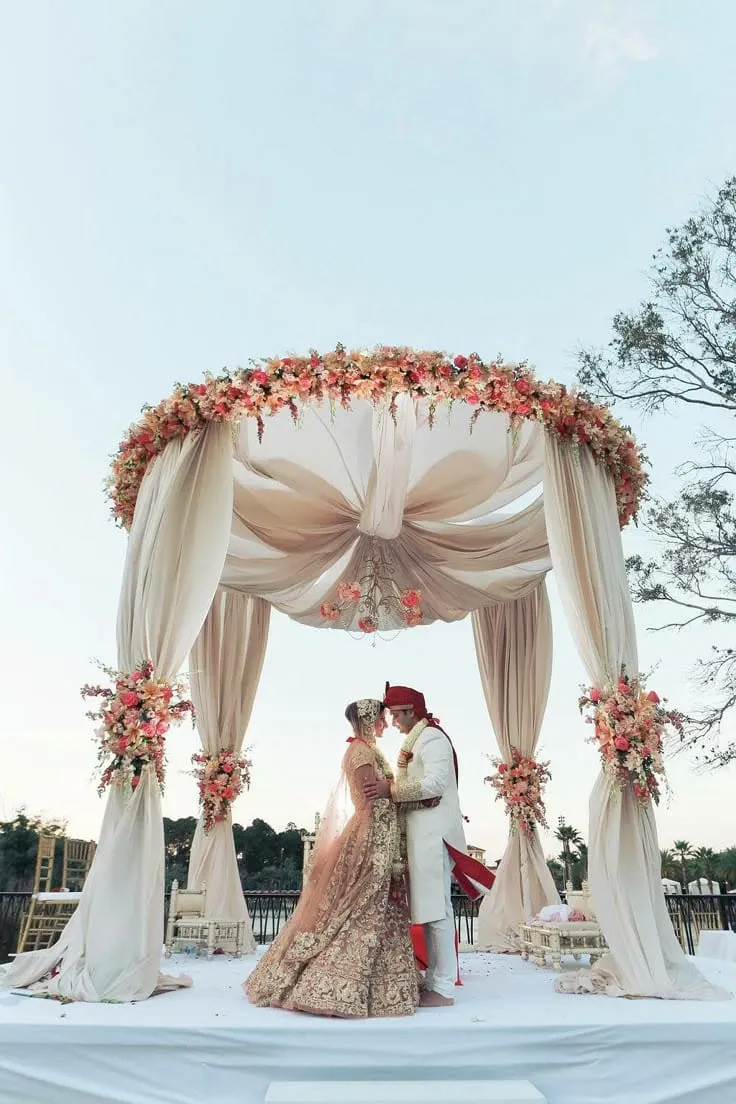 Bride and groom embracing under an ivory-draped floral mandap beside a lake
